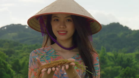 close-up-of-young-female-Vietnamese-farmer-standing-in-rice-field-holding-crop-from-harvesting-wearing-bamboo-hat-smiling-in-sow-motion-straight-to-camera