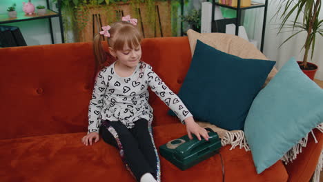 Smiling-young-children-girl-making-conversation-call-on-retro-phone-with-friends-on-couch-at-home