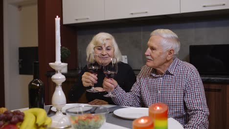 Portrait-of-senior-old-couple-toasting-wine-at-home-during-romantic-supper-in-kitchen-at-home