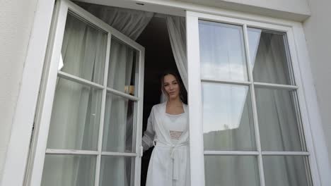 Bride-in-white-boudoir-dress-goes-to-balcony-and-opens-curtains-and-door-in-hotel-room-apartment