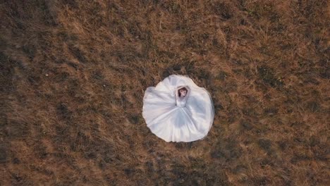 Beautiful-and-lovely-bride-in-wedding-dress-lie-on-grass-on-field.-Aerial-view