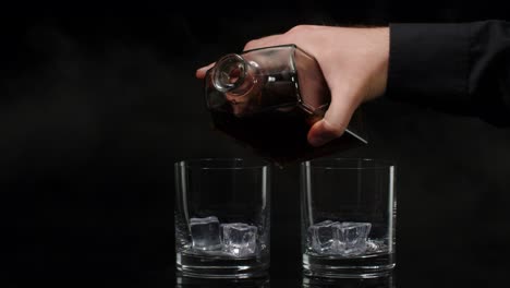 Barman-pouring-whiskey,-cognac,-brandy-from-bottle-into-glasses-with-ice-cubes-on-black-background