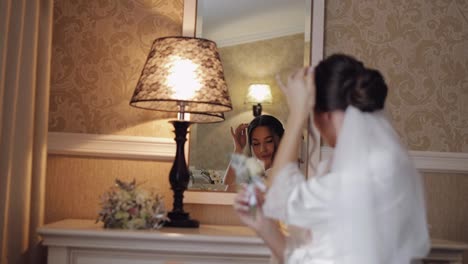 Bride-in-white-boudoir-dress-sitting-near-mirror-looking-at-reflection,-wedding-preparations-at-home