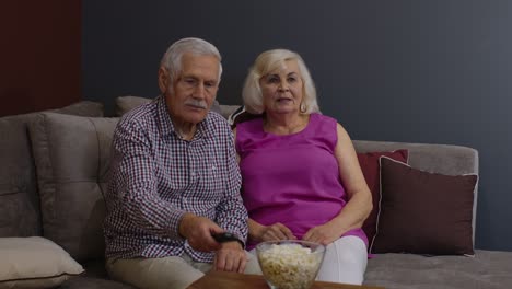 Senior-old-couple-holding-remote-control-talking-laughing-watching-humor-tv-show-sitting-on-sofa