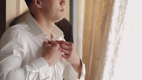 Un-Hombre-Elegante-Con-Chaqueta-Se-Abrocha-Los-Botones-Y-Se-Abrocha-La-Camisa-Blanca-Preparándose-Para-Salir-Cerca-De-La-Ventana