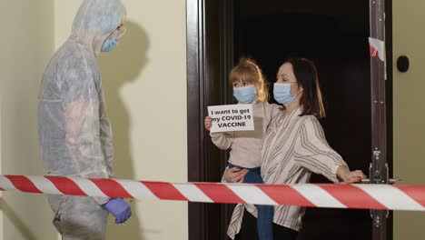 Doctor-in-protective-suit-with-vaccine-ampoule-and-syringe-visiting-mother-with-daughter-at-home