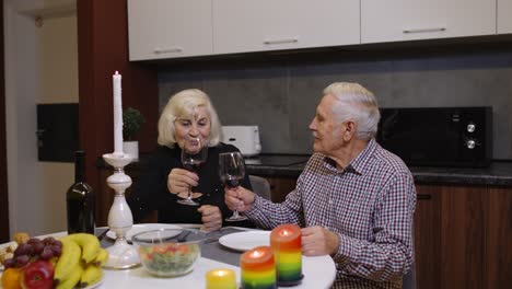 Portrait-of-senior-old-couple-toasting-wine-at-home-during-romantic-supper-in-kitchen-at-home