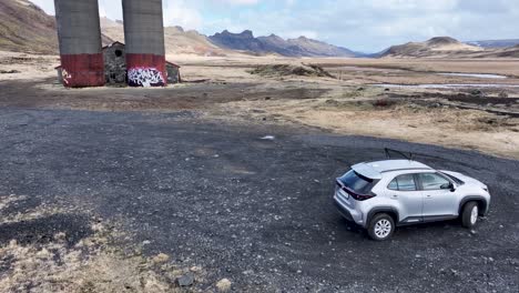 Drohnenaufnahmen-Zeigen-Eine-Verlassene-Farm-In-Island,-Umgeben-Von-Bergen,-Mit-Einem-Silbernen-Geländewagen,-Verfallenen-Gebäuden-Und-Lebhaften-Graffiti-Auf-Silos