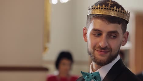 Groom-in-the-crown-stands-in-church.-He-looks-at-bride-and-smiles.-Newlyweds