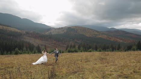 Lovely-young-newlyweds-bride-groom-running-on-mountain-slope,-holding-hands,-wedding-couple-family