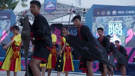 Hombres-Indonesios-Bailando-En-El-Campeonato-De-Surf-Profesional-Nias-En-Indonesia-Con-Escudos-Y-Lanzas-2023,-Mujeres-Indonesias-Con-Vestidos-Amarillos-Al-Fondo