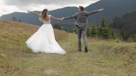 Lovely-young-newlyweds-bride-groom-running-on-mountain-slope,-holding-hands,-wedding-couple-family