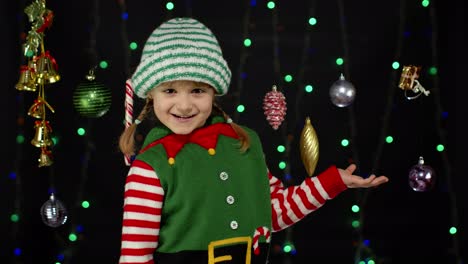 Kid-girl-in-Christmas-elf-Santa-helper-costume-point-with-hand-at-blank-space-shows-advertising-area