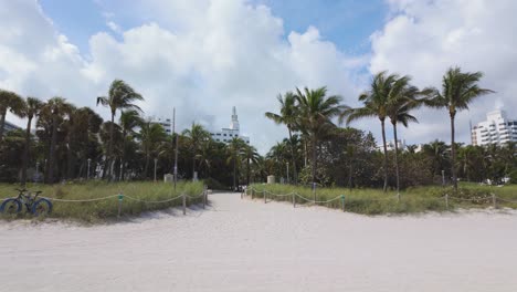 Pathway-leading-to-a-sandy-beach-with-palm-trees-and-buildings-in-the-background-in-Miami-Beach,-USA