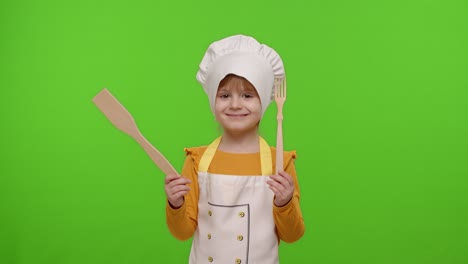 Child-girl-kid-dressed-as-cook-chef-showing-wooden-fork-and-spatula,-smiling,-nods-head-in-agreement