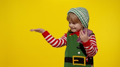 Kid-girl-in-Christmas-elf-Santa-helper-costume-points-fingers-at-blank-space-shows-advertising-area