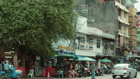 Busy-street-in-Bangkok's-Dusit-District-with-cars,-shops,-and-people-on-a-warm-day