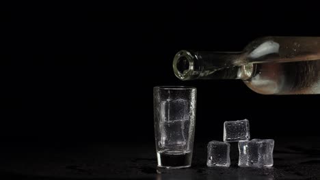 Bartender-pouring-up-shot-of-vodka-with-ice-cubes-from-bottle-into-shot-glass-on-black-background