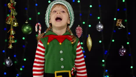 Teen-kid-girl-in-Christmas-elf-Santa-Claus-helper-costume-showing-thumbs-up-on-black-background