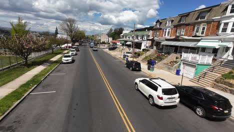 Vuelo-FPV-Sobre-Una-Calle-En-Un-Suburbio-Estadounidense-Con-Autos-En-Movimiento-Y-Una-Hilera-De-Casas-En-Un-Día-Soleado