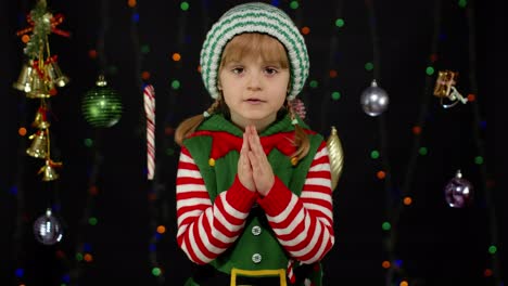 Kid-in-Christmas-elf-Santa-helper-costume-begs,-prays-about-gifts-on-New-Year.-Keeps-palms-together