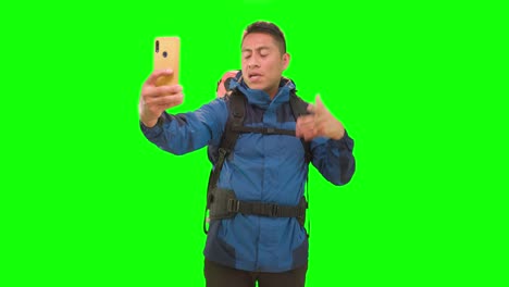 Man-with-hiking-backpack-and-yellow-smartphone-in-hand-in-a-studio-with-green-screen,-recording-content-as-a-travel-influencer