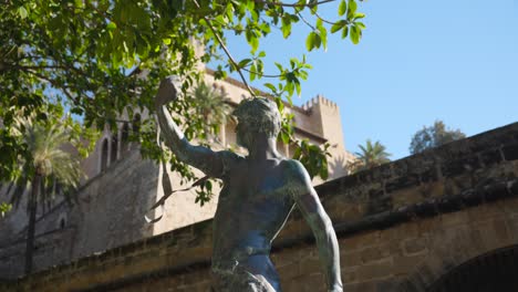 La-Estatua-De-Bronce-Del-Hondero-De-Lorenzo-Rosselló,-Situada-En-Mallorca,-Con-Una-Arquitectura-Histórica-De-Piedra-Y-Vegetación-Al-Fondo