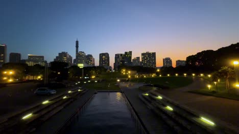 City-park-at-dusk-with-illuminated-pathways-and-skyline-in-the-background,-aerial-view