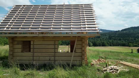 Wooden-house-under-construction,-made-of-logs-and-located-in-beautiful-landscape-during-a-beautiful-summer-day,-surrounded-by-lush-greenery,-grass-and-trees-under-a-blue-sky