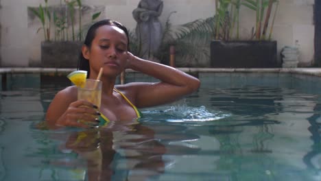 Toma-En-Cámara-Lenta-De-Una-Mujer-Asiática-Con-Un-Bikini-Verde-Y-Amarillo-Disfrutando-De-Un-Refrescante-Cóctel-Tropical-En-La-Piscina.