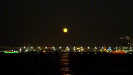 Full-moon-rises-over-a-city-harbor-with-colorful-boats-and-sparkling-lights-reflecting-on-the-water