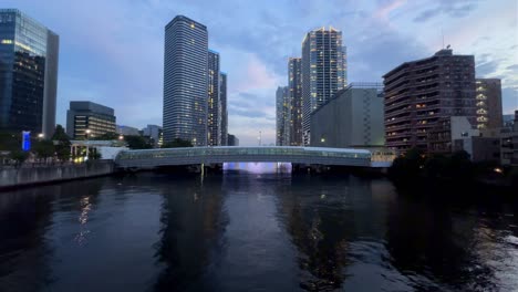 Bridge-over-calm-river-at-dusk-with-cityscape-background-reflecting-in-the-water