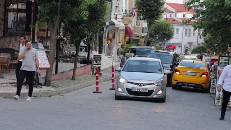 Car-traffic-on-the-narrow-streets-of-Istanbul,-Turkey,-during-the-daytime,-with-vehicles-driving-near-pedestrian-areas-featuring-cafes-and-restaurants