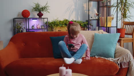 Child-toddler-hold-smartphone-watching-funny-cartoons,-chatting-with-friends-on-couch-alone-at-home