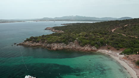 Una-Tranquila-Cala-De-Cerdeña-Con-Barcos-Anclados-Y-Exuberante-Vegetación,-Vista-Aérea