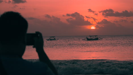 Der-Mann-In-Der-Silhouette-Fotografiert-Den-Majestätischen,-Brennenden-Sonnenuntergang-über-Dem-Meer-An-Der-Strandlandschaft-Von-Jimbaran-Mit-Fischerbooten,-Die-In-Ufernähe-Vor-Anker-Liegen