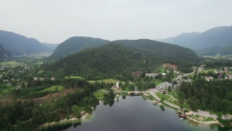 Toma-Aérea-Con-Dron-Sobre-El-Lago-Bohinj-En-Eslovenia-Con-Una-Iglesia-Y-Montañas-De-Fondo-Mientras-Se-Pone-El-Sol