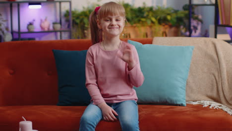 Child-girl-sitting-on-sofa-at-home-looking-at-camera-smiling-waving-hands-gesturing-hello-or-goodbye