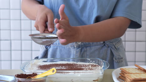 Front-View-Of-Woman's-Hand-Sprinkles-Cocoa-On-Tiramisu-Cake-In-A-Glass-Bowl-At-Kitchen