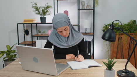 Muslim-business-woman-study-with-notebook-at-home-office-on-laptop-computer-talking-to-webcam-online