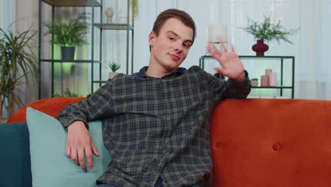 Teenager-man-smiling-friendly-at-camera,-waving-hands-gesturing-hello,-hi,-greeting-at-home-on-couch