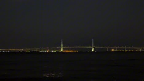 Illuminated-bridge-stretches-over-dark-water-with-city-lights-in-the-background