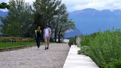 Traveling-Couple-Holding-Hands-While-Walking-With-Mountain-In-Background