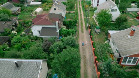 Two-men-walk-along-dirt-road-between-summer-houses-in-lush-green-area-next-to-a-river