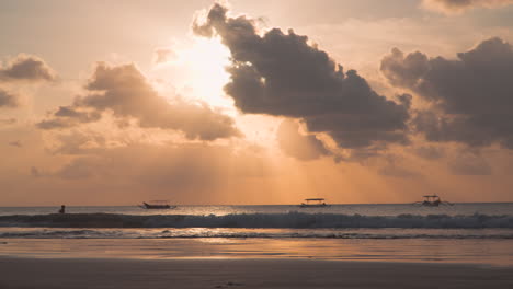 Un-Hombre-Silueteado-Entra-Al-Agua-Del-Mar-A-Contraluz-Con-El-Sol-Dorado-Poniéndose-Sobre-El-Horizonte,-Un-árbol,-Viejos-Barcos-Pesqueros-Balineses-Amarrados-Flotando