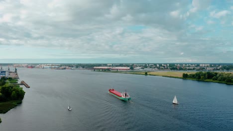 Aerial-view-of-cargo-ship-sailing-through-river-channel-along-harbor-with-two-smaller-sailboats-on-each-side