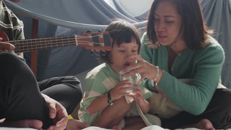 Glückliche-Kleine-Asiatische-Tochter-Trinkt-Wasser-Aus-Einer-Tasse,-Während-Mutter-Ihr-Hilft-Und-Vater-Mit-Der-Gitarre-In-Der-Nähe-Sitzt