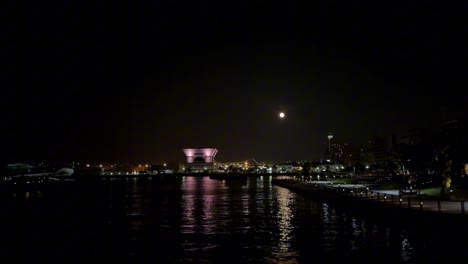 Stadtufer-Bei-Nacht-Mit-Beleuchteten-Gebäuden-Und-Spiegelungen-Auf-Dem-Wasser-Unter-Einem-Mondbeschienenen-Himmel