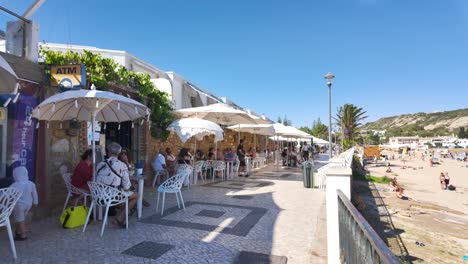 A-sunny-day-scene-showing-a-charming-coastal-café-with-outdoor-seating-next-to-a-sandy-beach,-where-people-enjoy-their-time-in-Praia-da-Luz