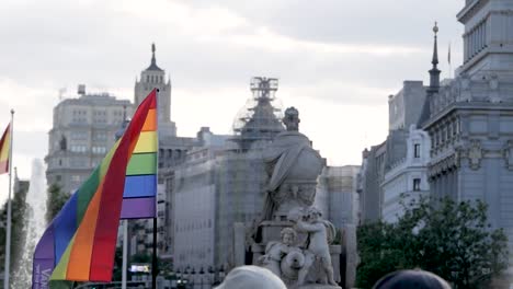 Primer-Plano-De-Una-Bandera-Del-Arcoíris-Y-Banderas-Españolas-Cerca-De-Una-Estatua,-Que-Simbolizan-El-Orgullo-Y-El-Patrimonio-Nacional